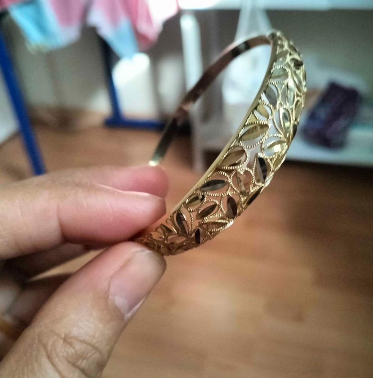 Dłoń trzyma złoty, ażurowy mankiet bransoletki z motywem liści na rozmytym tle domowego wnętrza.
