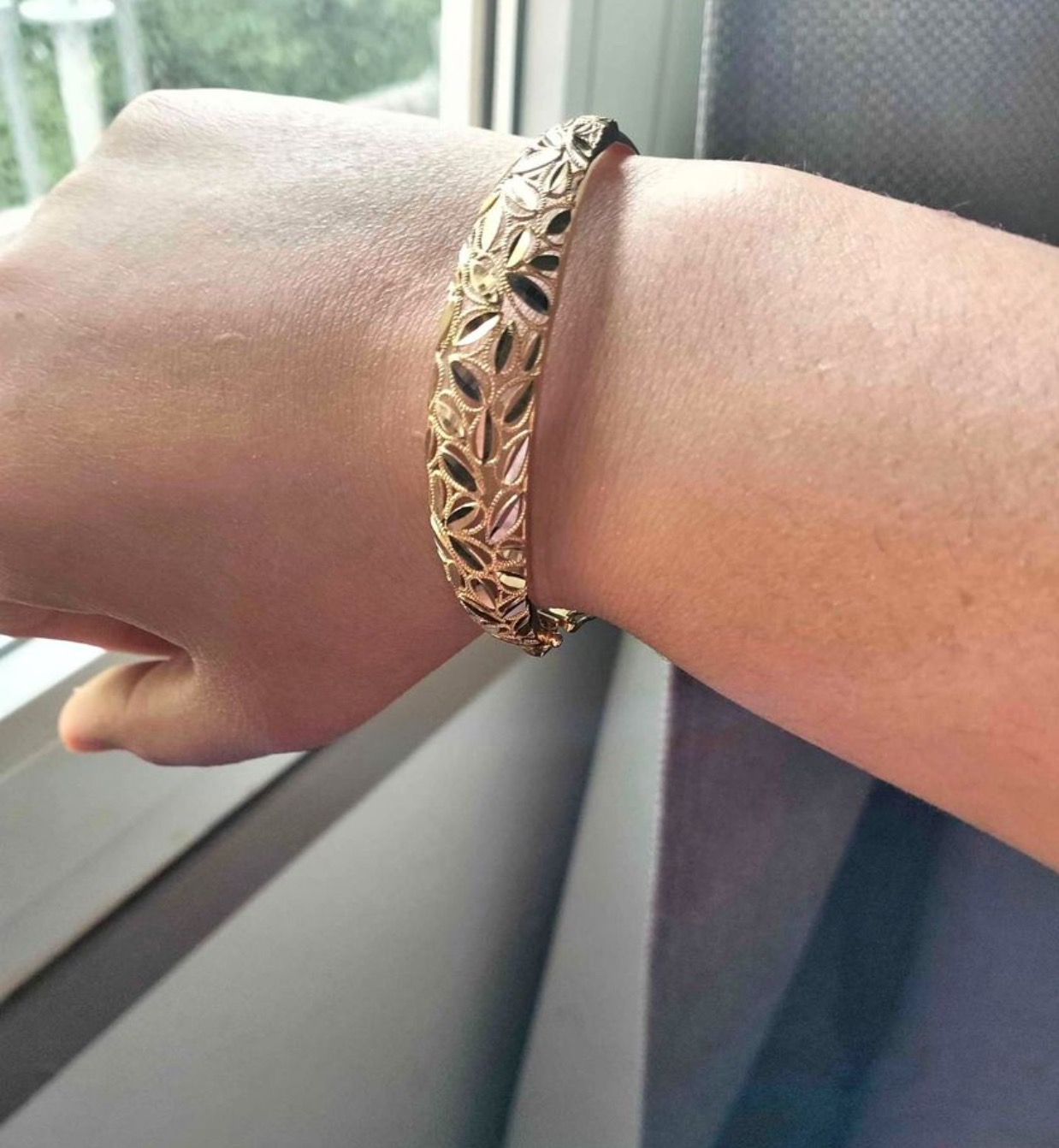 Złota bransoletka z ażurowym wzorem liści na nadgarstku, widoczna w naturalnym świetle padającym z okna.
