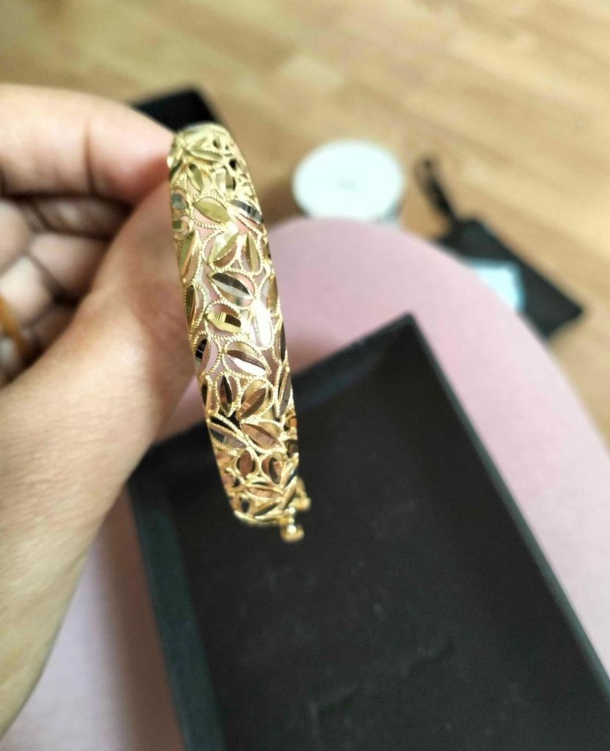 Dłoń prezentuje złoty, ażurowy mankiet bransoletki z motywem liści na tle czarnego pudełka i różowego ubrania.
