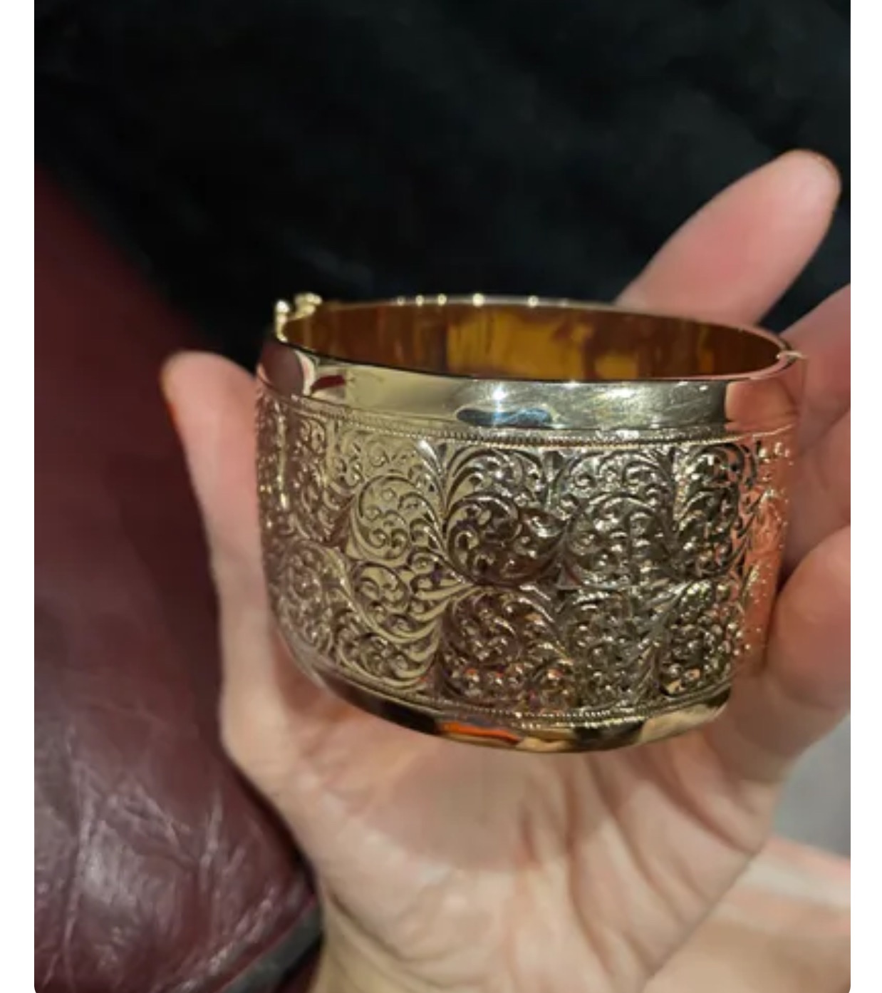 Szeroka, złota bransoletka z filigranowym wzorem kwiatowym, trzymana w dłoni na tle ciemnego materiału.