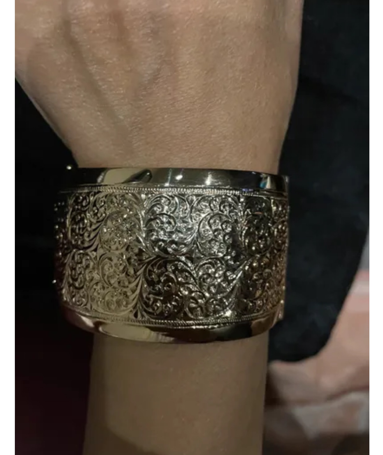 Złota, szeroka bransoleta z filigranowym, roślinnym wzorem na nadgarstku, w ciepłym oświetleniu.