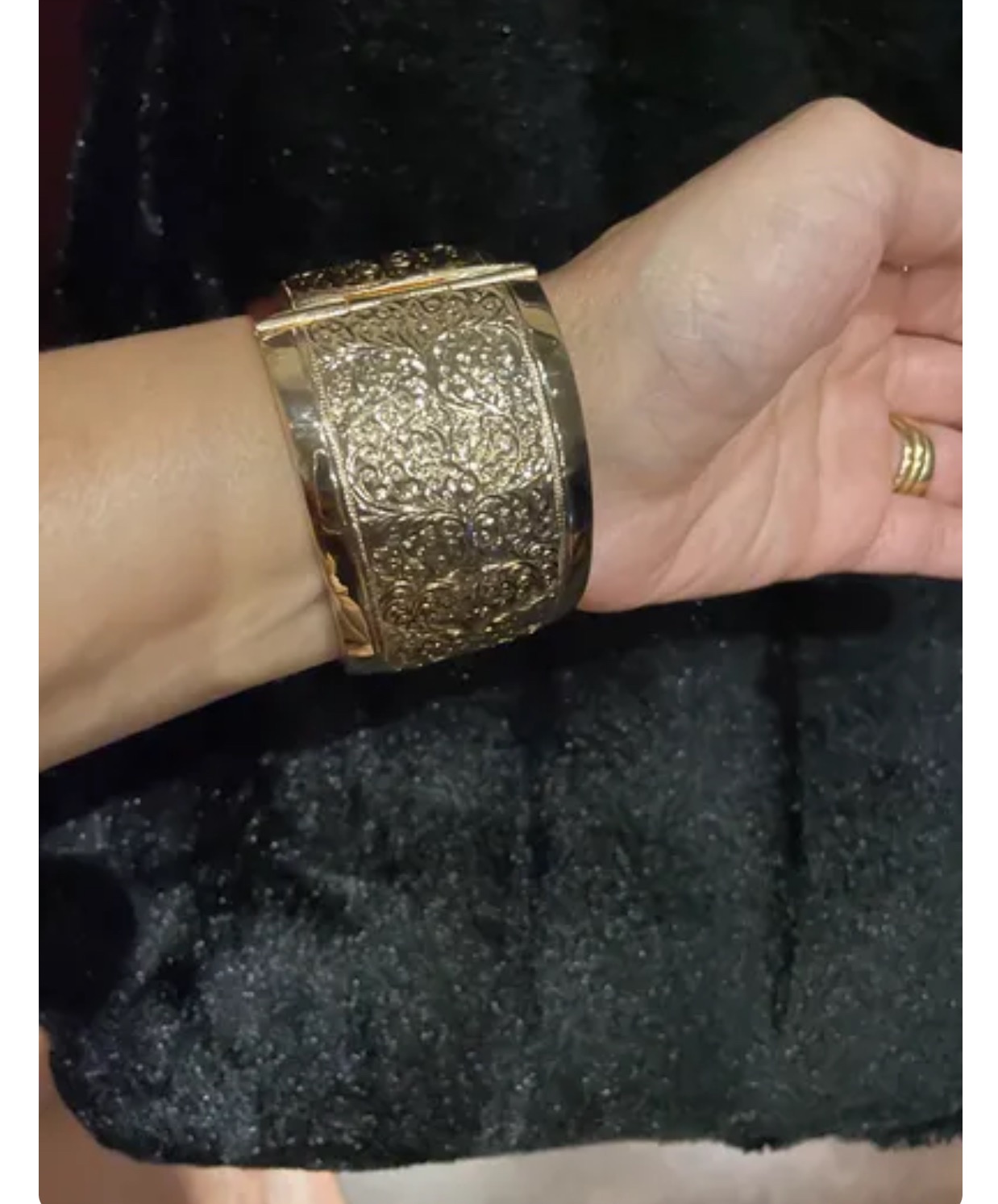 Złota, szeroka bransoleta z wytłaczanym, roślinnym wzorem na kobiecym nadgarstku, w tle czarny, błyszczący materiał.