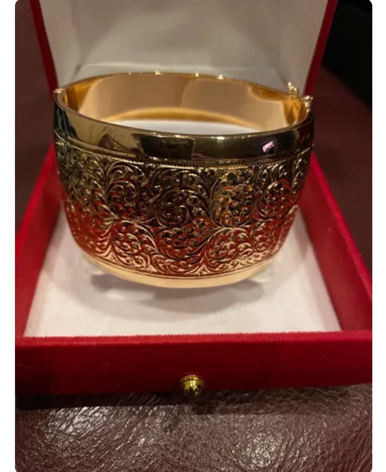 Złota, szeroka bransoleta z filigranowym, roślinnym wzorem, umieszczona w czerwonym pudełku z weluru na brązowym tle.