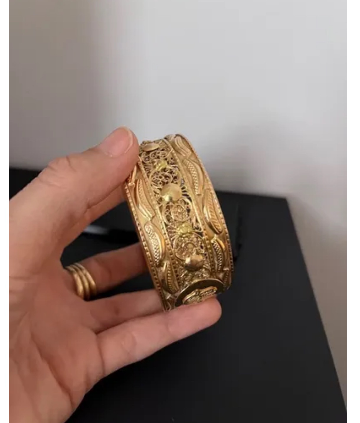 Dłoń prezentuje bogato zdobioną, ażurową, złotą bransoletę na czarnym tle.