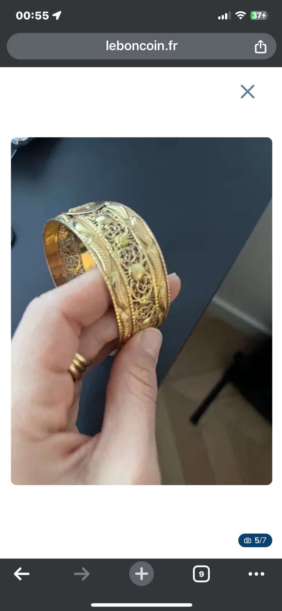 Dłoń z pierścionkiem trzymająca zdobioną, złotą bransoletę na czarnym tle.