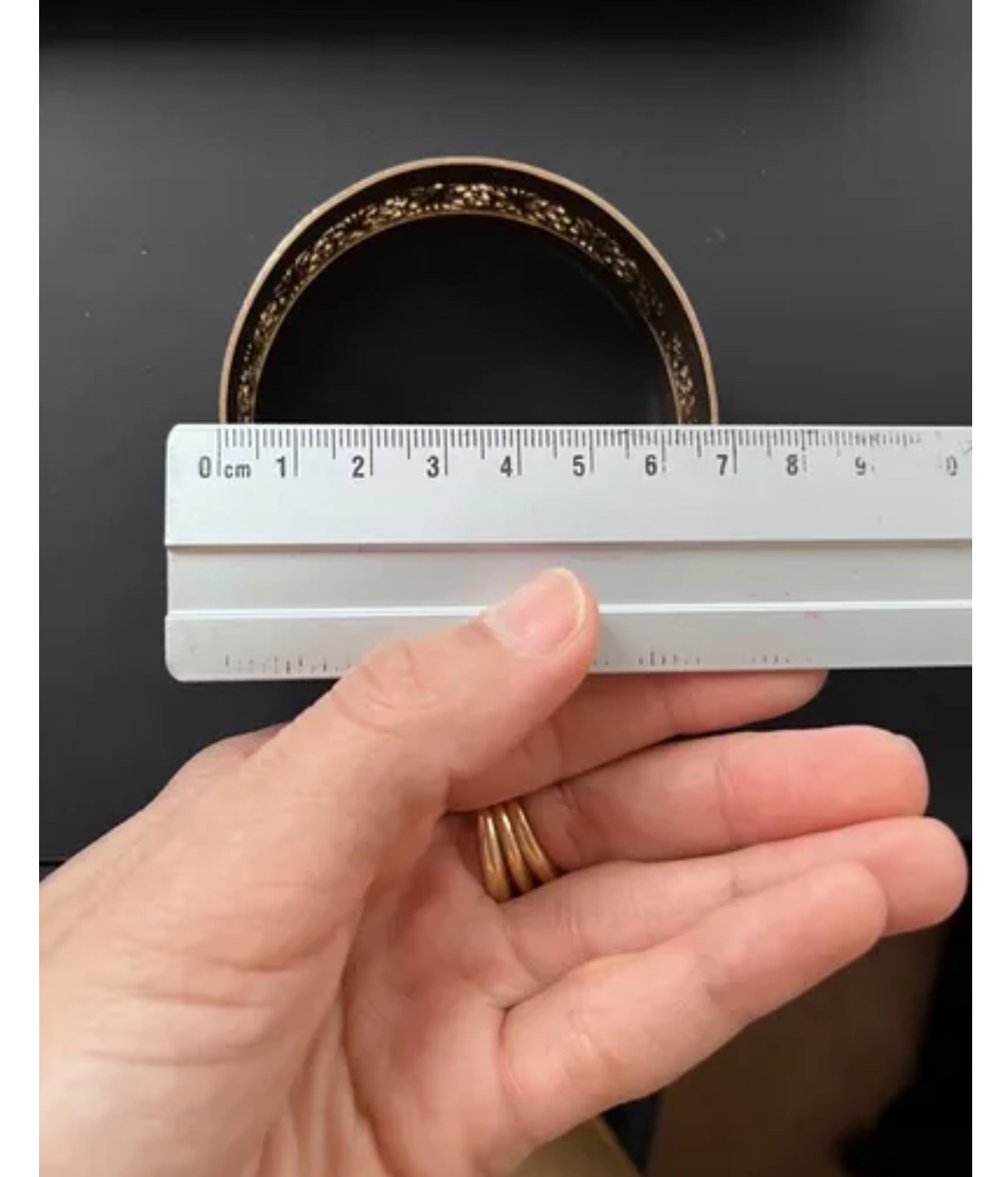 Pomiar szerokości obręczy z ozdobnym brzegiem za pomocą linijki trzymanej w dłoni z trzema złotymi pierścionkami.