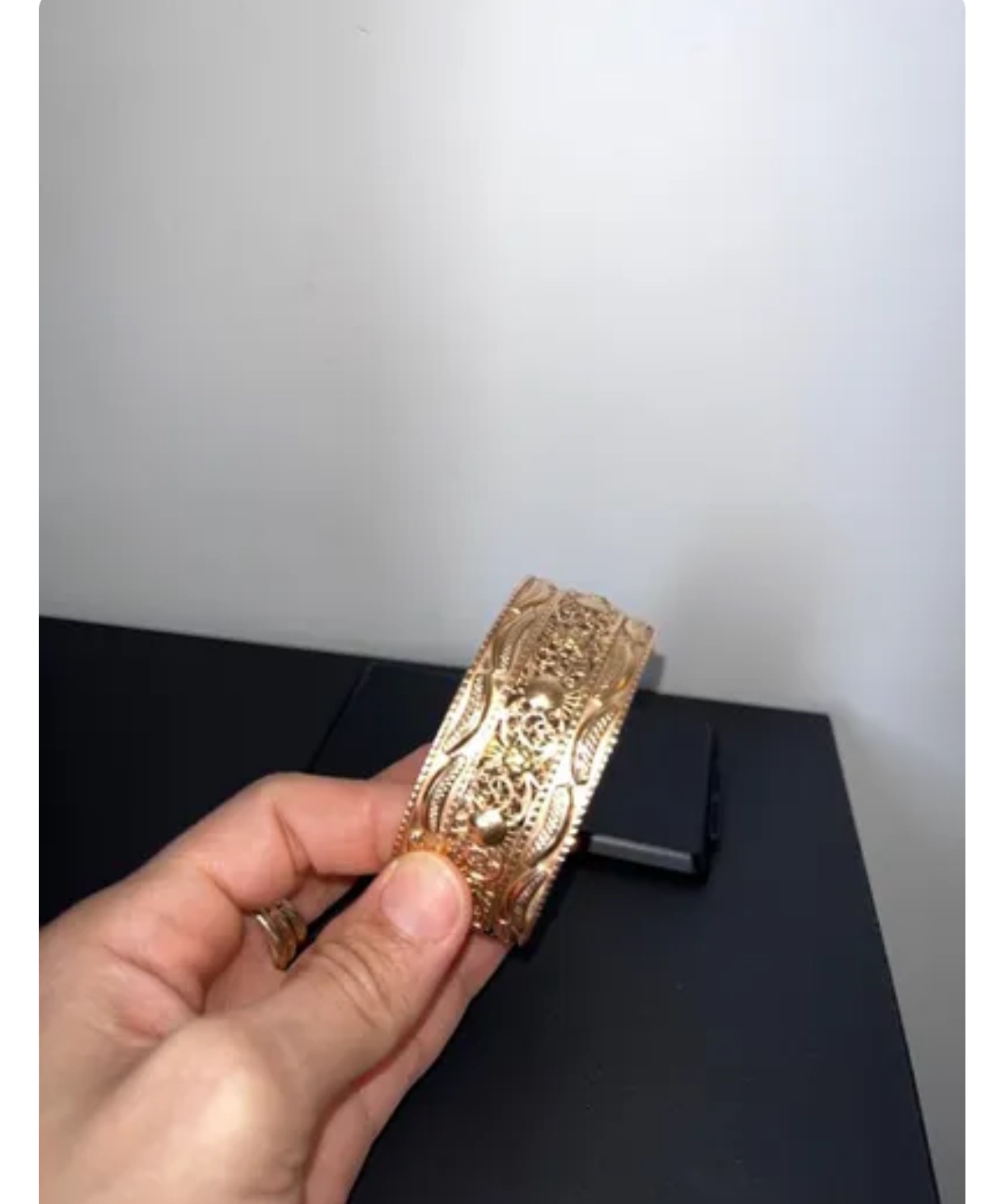 Dłoń kobiety prezentuje złoty, ażurowy mankiet bransolety na tle czarnego blatu.