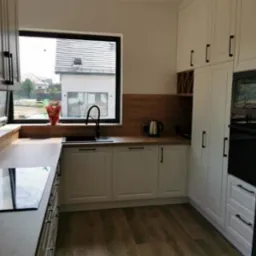 Jasna kuchnia z białymi szafkami, drewnianym blatem i czarnymi akcentami, widok na ogr&oacute;d przez okno, minimalistyczny design.