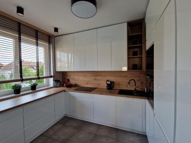 Jasna kuchnia z białymi, lakierowanymi frontami szafek, drewnianym blatem i okładziną ścienną, żaluzjami w oknie, oświetleniem punktowym i otwartą półką z książkami.