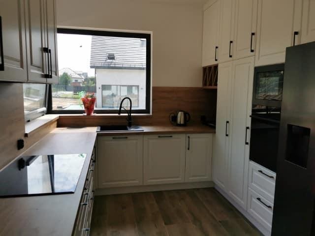 Jasna kuchnia z białymi szafkami, drewnianym blatem i czarnymi akcentami, widok na ogród przez okno, minimalistyczny design.