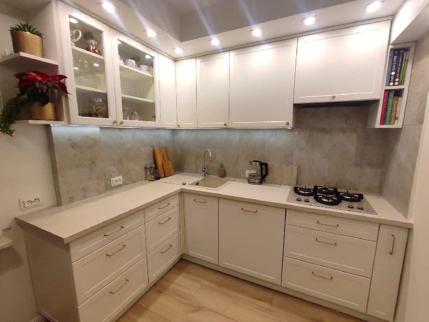 Jasna kuchnia z białymi frontami, przeszklonymi szafkami wiszącymi, drewnianym blatem i płytą gazową, oświetlona punktowymi lampami sufitowymi.