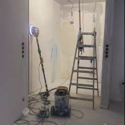 Rusbud - Wnętrze w trakcie prac przygotowawczych do malowania: szpachlowane ściany, szlifierka do gipsu z odkurzaczem, drabina i lampa budowlana.