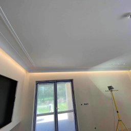 Rusbud - Świeżo wykończone wnętrze z oświetleniem LED w listwach sufitowych, okno zabezpieczone folią malarską, widoczny reflektor na statywie.