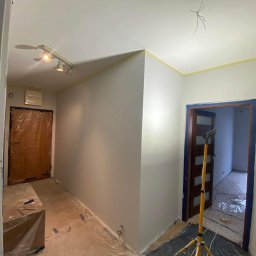 Rusbud - Korytarz w trakcie odświeżania: świeżo pomalowane ściany w odcieniach szarości, zabezpieczone folią podłogi, taśma malarska przy suficie, widoczna lampa budowlana i zakryte folią drzwi.