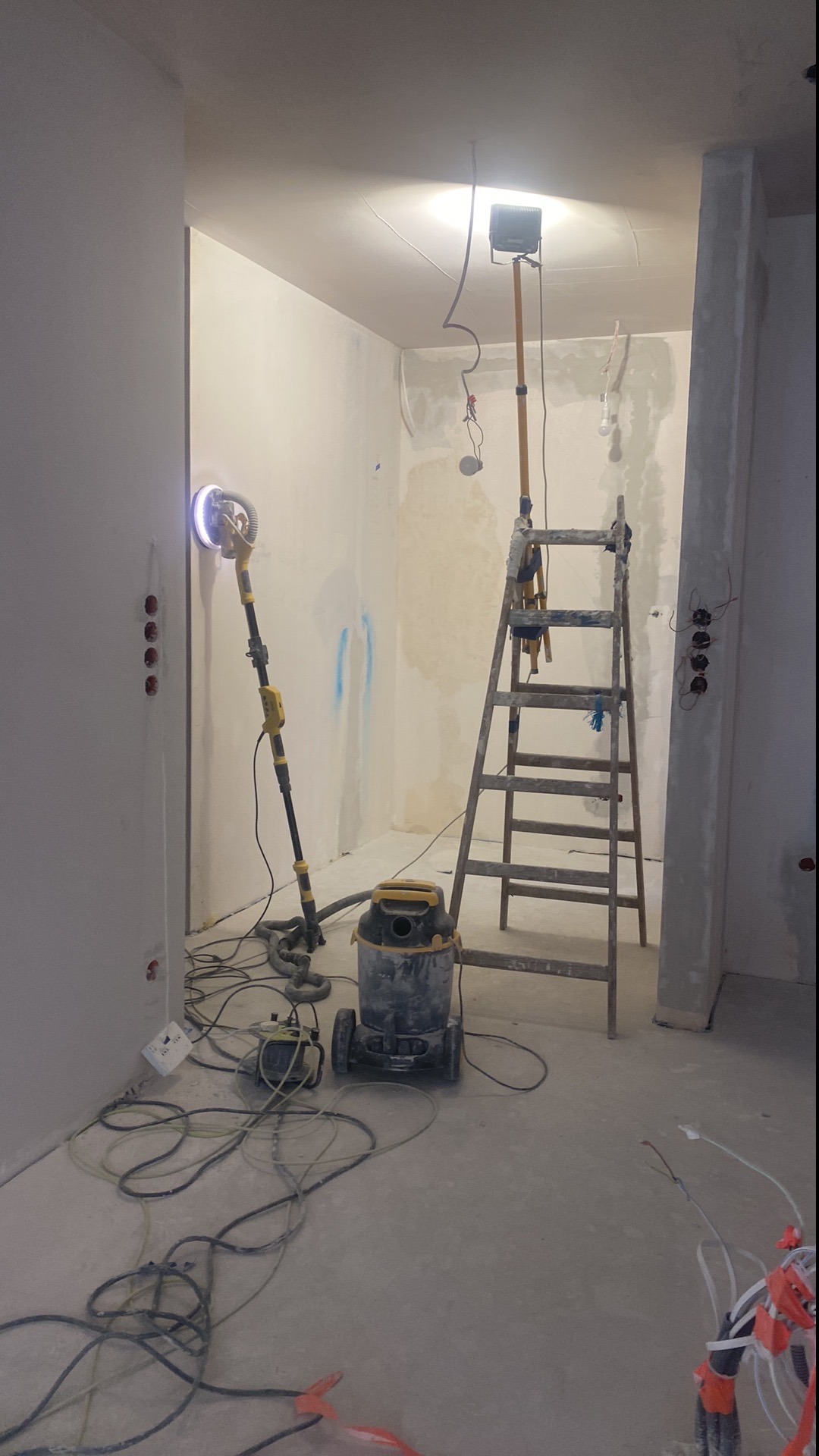 Wnętrze w trakcie prac przygotowawczych do malowania: szpachlowane ściany, szlifierka do gipsu z odkurzaczem, drabina i lampa budowlana.