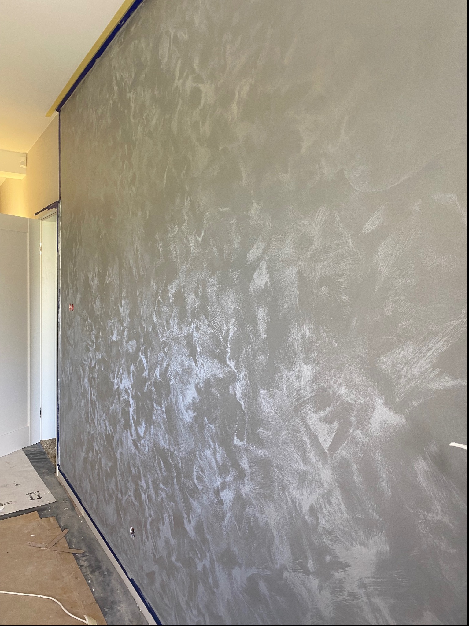 Ściana z dekoracyjną, szarą strukturą, zabezpieczona taśmą malarską przy suficie i podłodze, widoczne gniazdko elektryczne i fragment podłogi z kartonami ochronnymi.