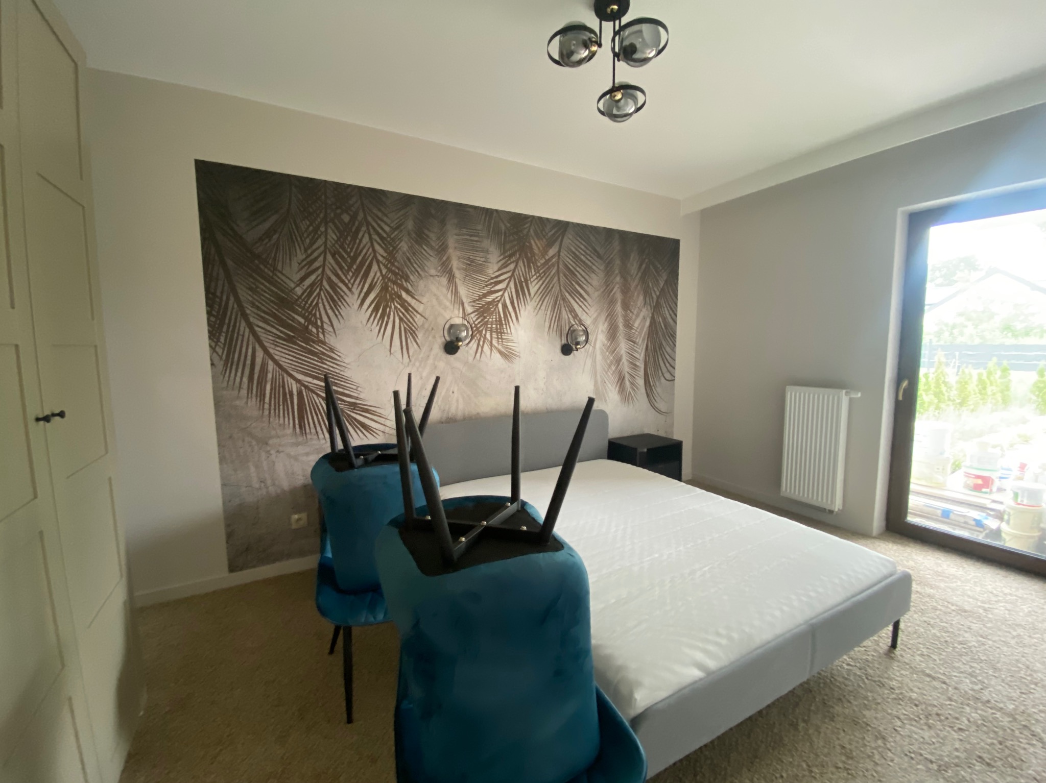 Sypialnia po remoncie z łóżkiem, szafką nocną i fototapetą z motywem palm, dwa krzesła odwrócone do góry nogami, widoczny grzejnik i okno z widokiem na zieleń.