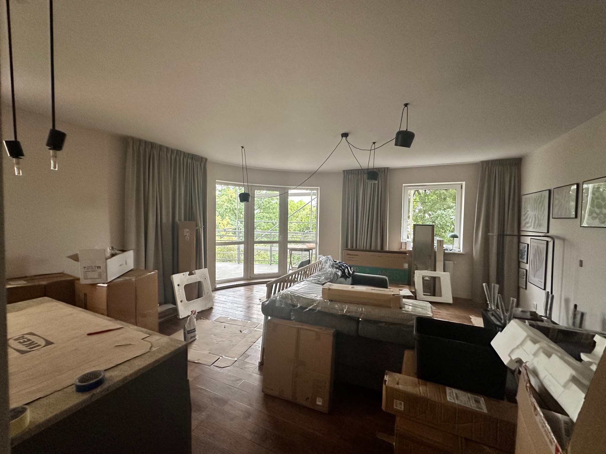 Pokój w trakcie przeprowadzki lub remontu: meble zabezpieczone folią, kartony porozstawiane, widoczne ślady po pracach wykończeniowych na podłodze, okna z widokiem na zieleń.