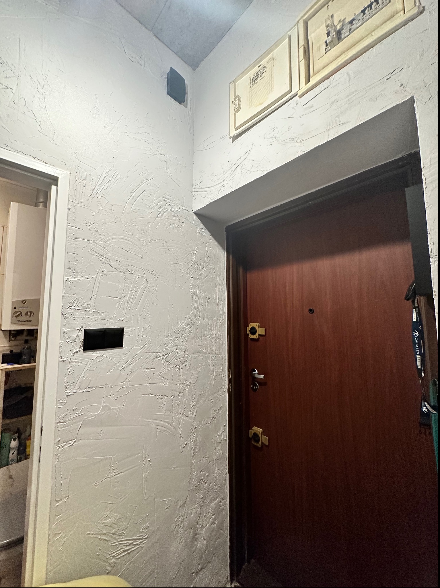 Korytarz w Suwałkach po remoncie: ściany pokryte białą strukturą, brązowe drzwi wejściowe, widoczne detale wykończenia i czarny włącznik światła na ścianie.
