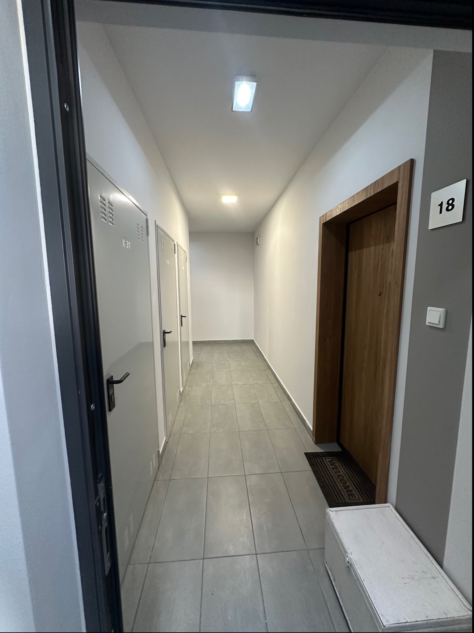 Korytarz w budynku mieszkalnym z szarymi płytkami na podłodze, rzędem białych drzwi do komórek lokatorskich po lewej i drewnianymi drzwiami do mieszkania nr 18 po prawej stronie.