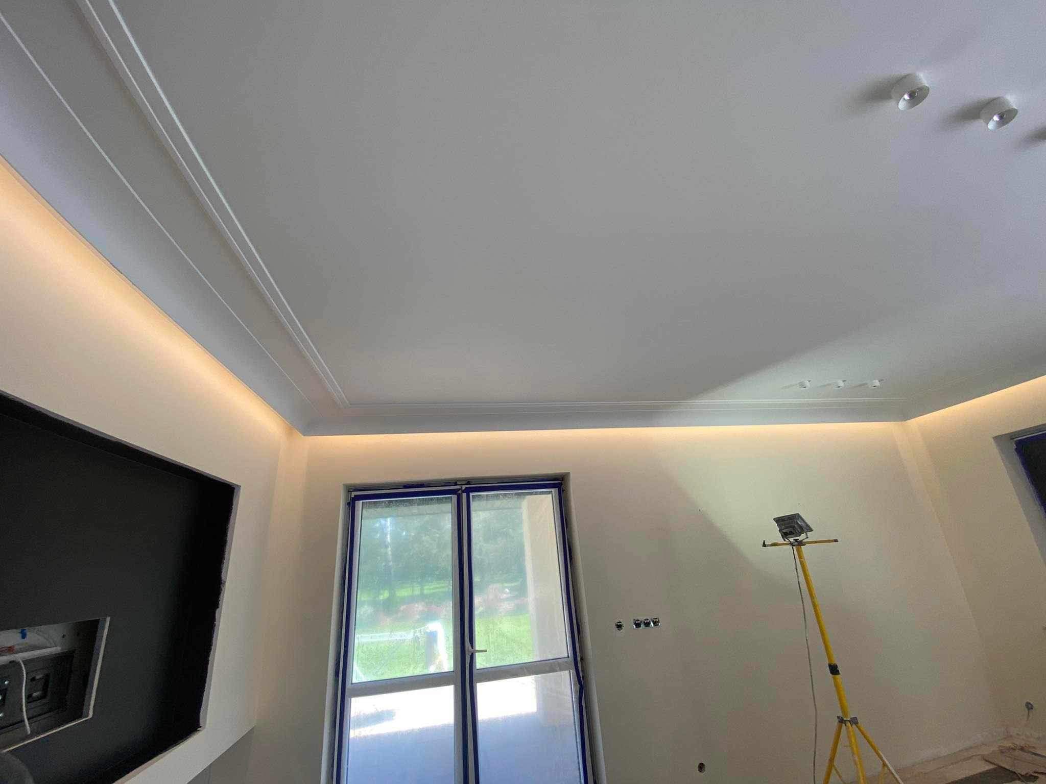 Świeżo wykończone wnętrze z oświetleniem LED w listwach sufitowych, okno zabezpieczone folią malarską, widoczny reflektor na statywie.