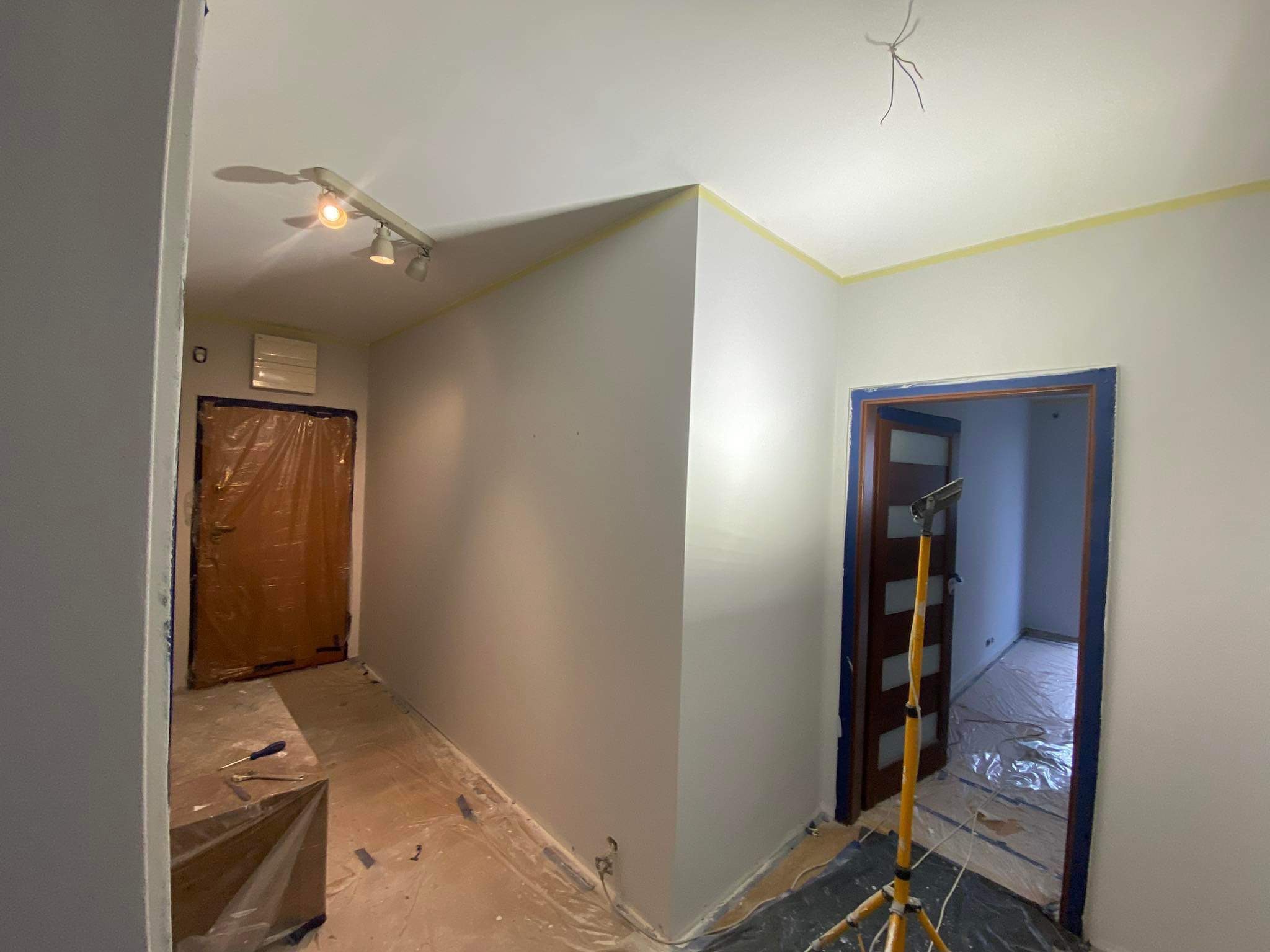 Korytarz w trakcie odświeżania: świeżo pomalowane ściany w odcieniach szarości, zabezpieczone folią podłogi, taśma malarska przy suficie, widoczna lampa budowlana i zakryte folią drzwi.