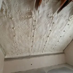 Poddasze z drewnianą konstrukcją pokryte warstwą pianki poliuretanowej, widoczne punkty mocowania, surowe ściany w tle.