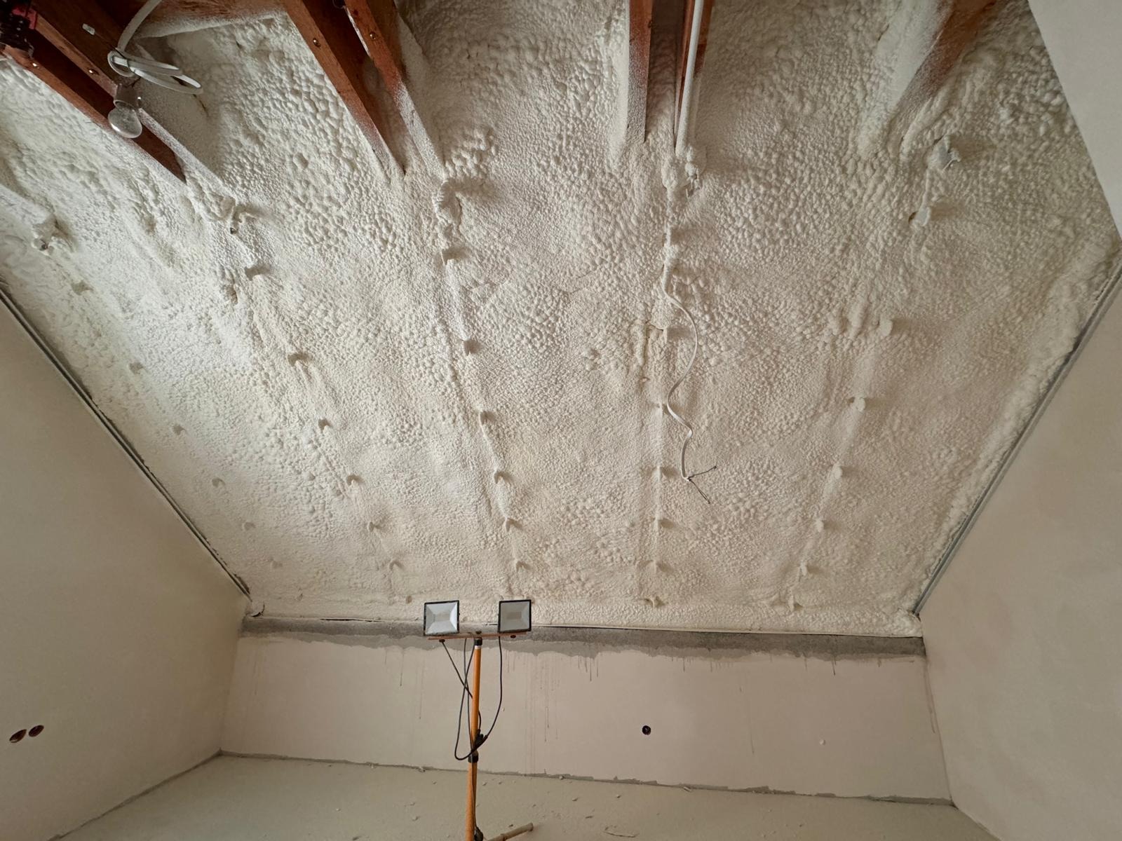 Poddasze po ociepleniu pianką poliuretanową, widoczne drewniane elementy konstrukcyjne i równomierna warstwa izolacji, na podłodze ustawione lampy budowlane.