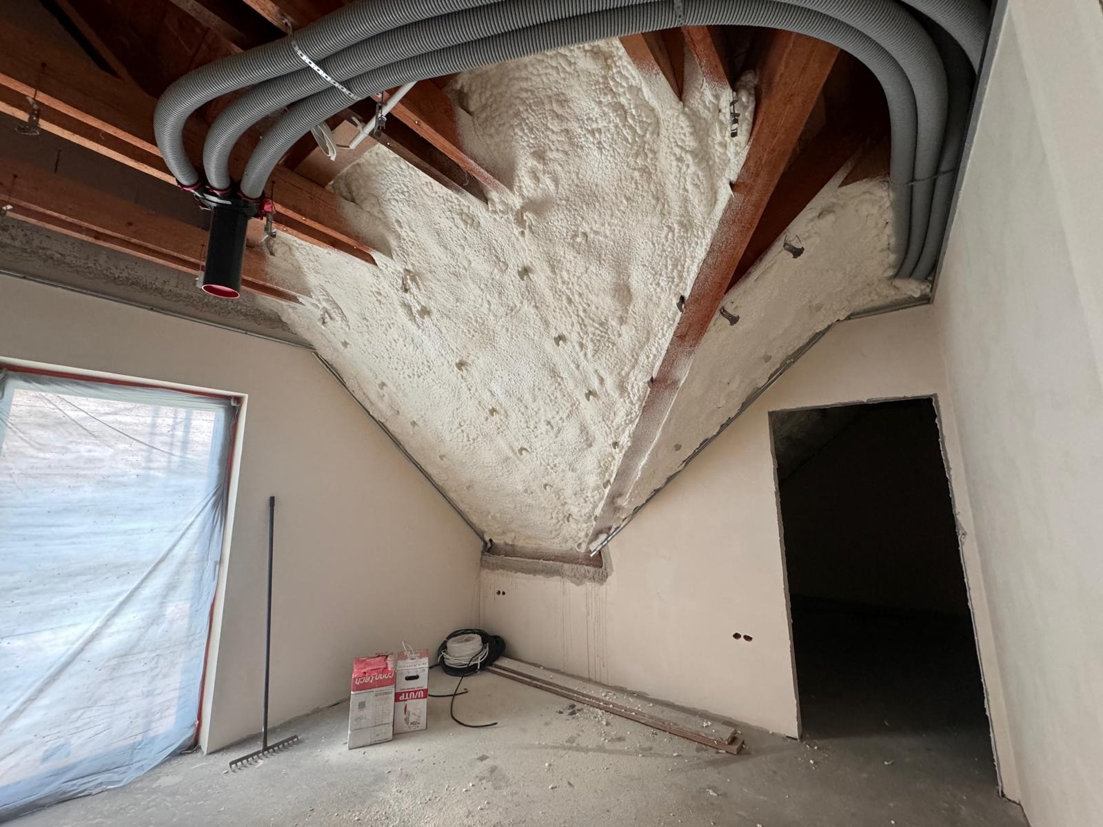 Poddasze po ociepleniu pianką PUR, widoczne drewniane krokwie i szare rury wentylacyjne pod sufitem, w tle okno zabezpieczone folią i drzwi do innego pomieszczenia, na podłodze leżą materiały...