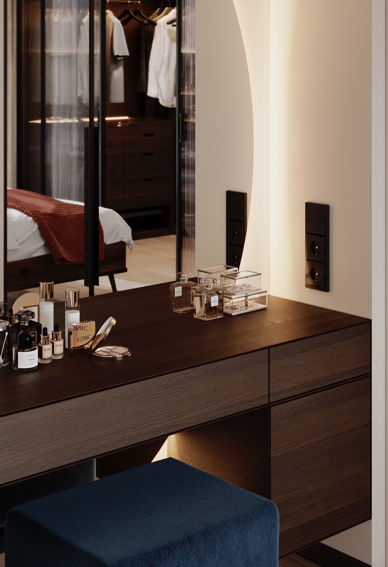 Toaletka z ciemnego drewna z kosmetykami i lustrem z podświetleniem, w tle szafa z ubraniami i łóżko z czerwoną narzutą.