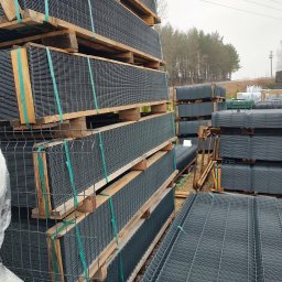SIATGOLD - Stosy paneli ogrodzeniowych zgrzewanych, zabezpieczonych taśmami na drewnianych paletach, składowane na zewnątrz w pochmurny dzień.