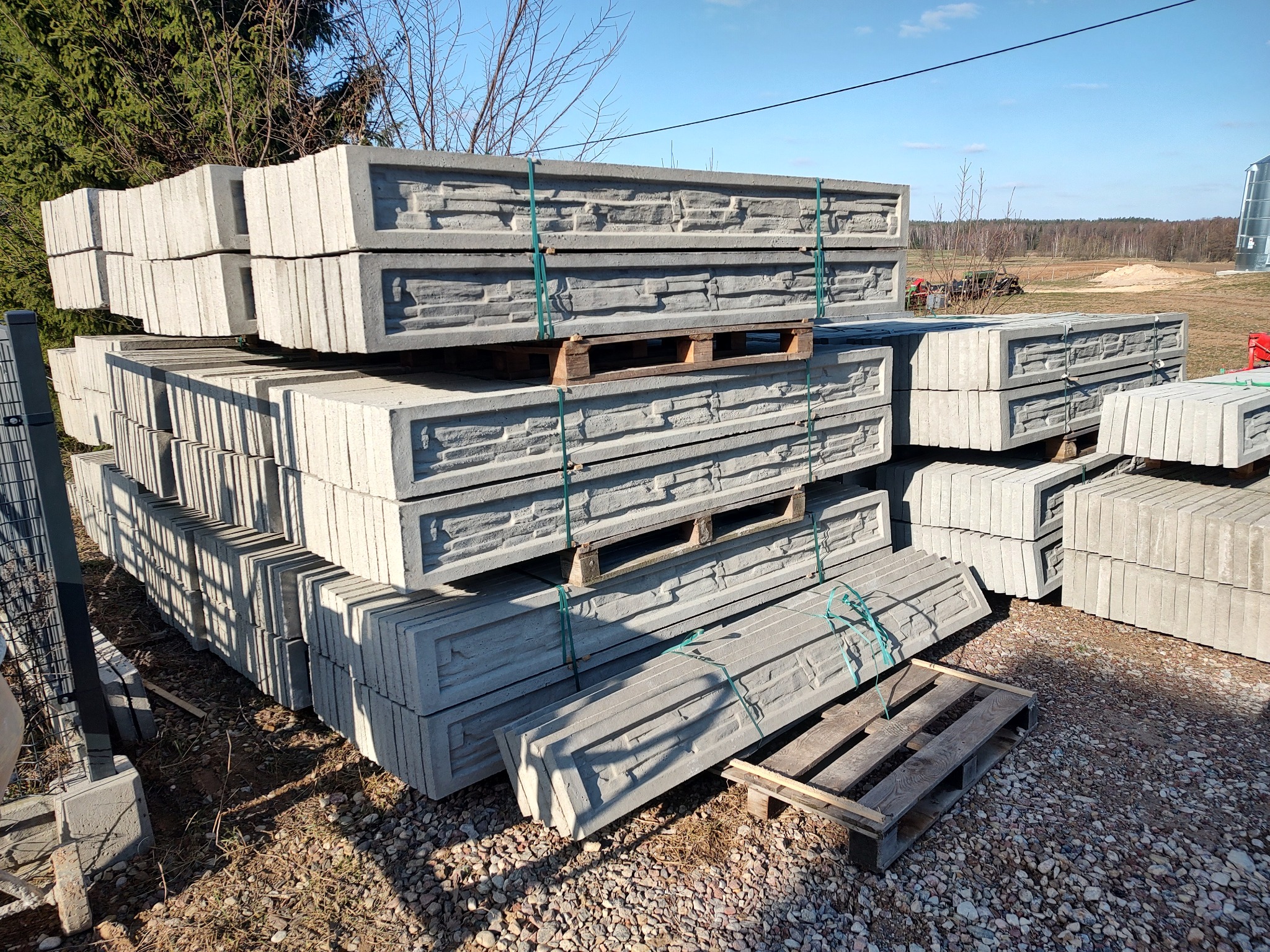 Stosy prefabrykowanych, betonowych elementów ogrodzeniowych z dekoracyjnym wzorem, spięte taśmami na drewnianych paletach, składowane na żwirowym podłożu w terenie niezabudowanym.