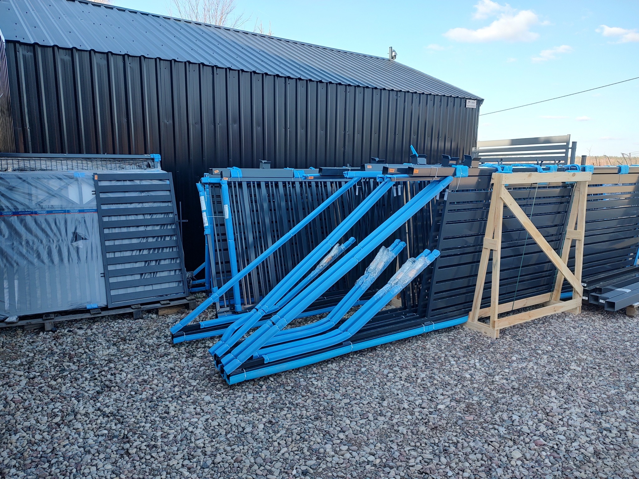 Stosy elementów ogrodzeniowych: panele, słupki z niebieskim zabezpieczeniem, ułożone na żwirowym podłożu przed budynkiem o ciemnej, blaszanej elewacji.