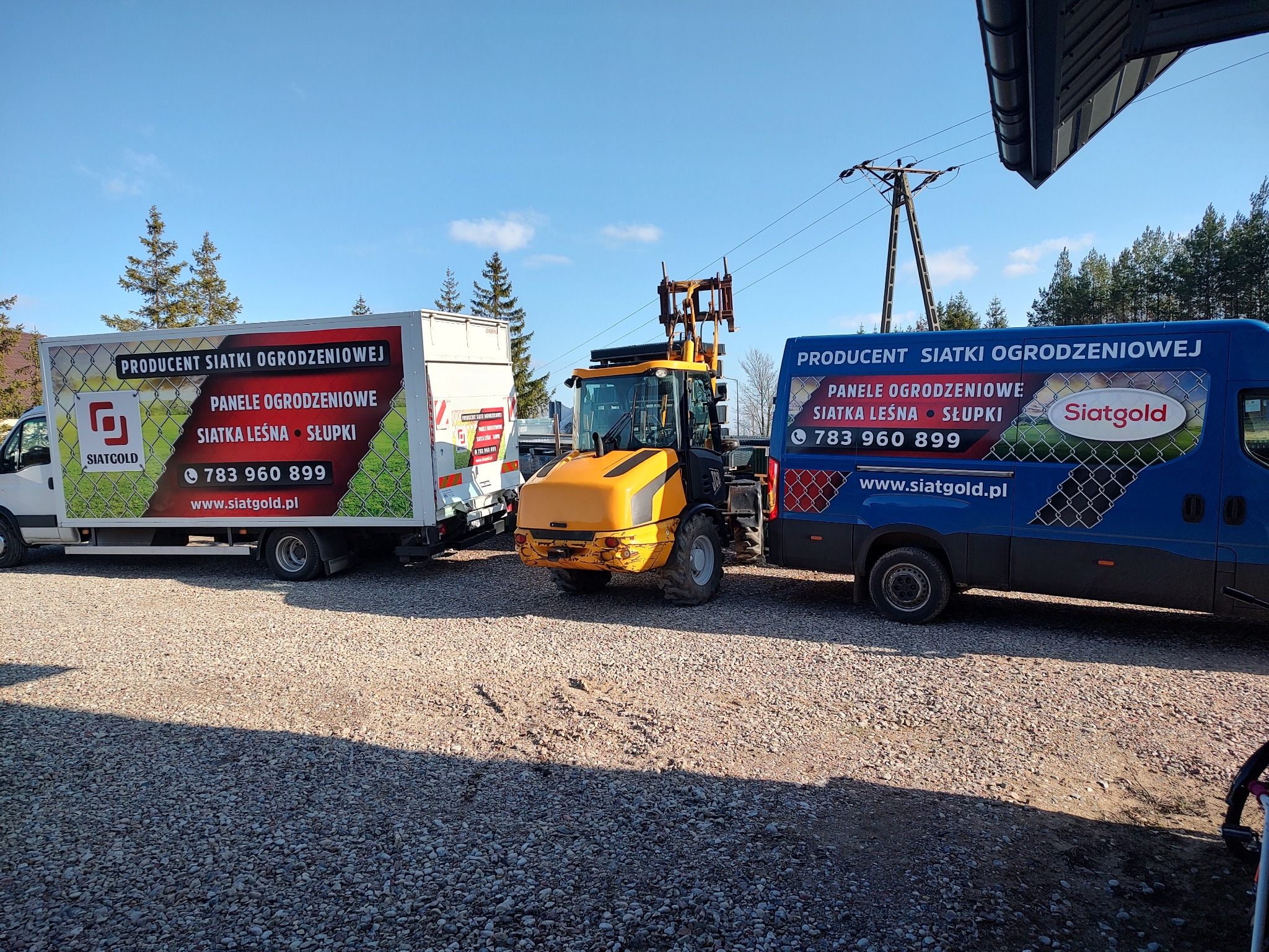 Dwa samochody dostawcze z reklamą producenta siatki ogrodzeniowej i paneli ogrodzeniowych, oraz żółta ładowarka teleskopowa na żwirowym parkingu w słoneczny dzień.