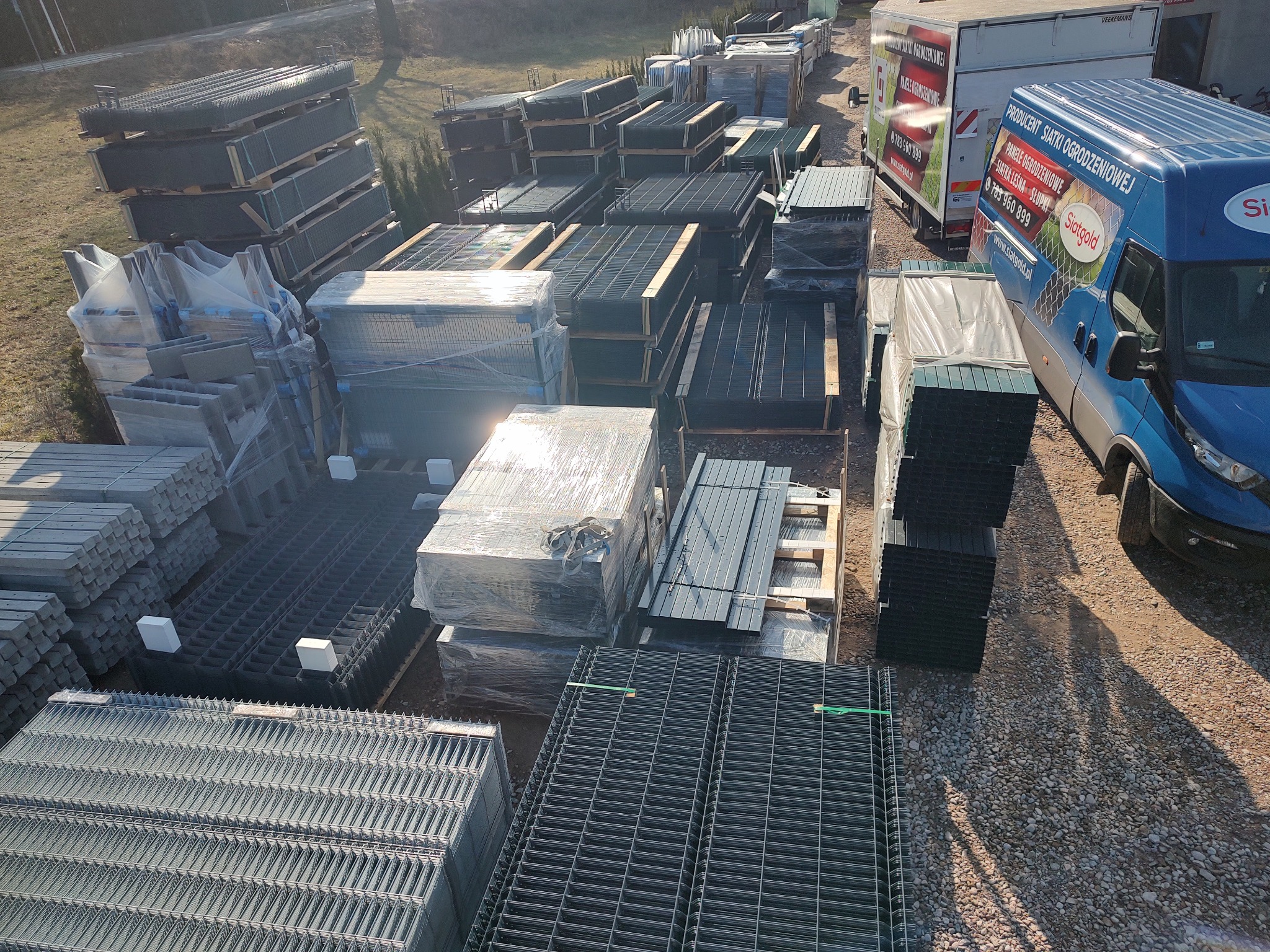 Składowisko elementów ogrodzeniowych: panele, słupki, siatka, z widocznymi samochodami dostawczymi w tle.