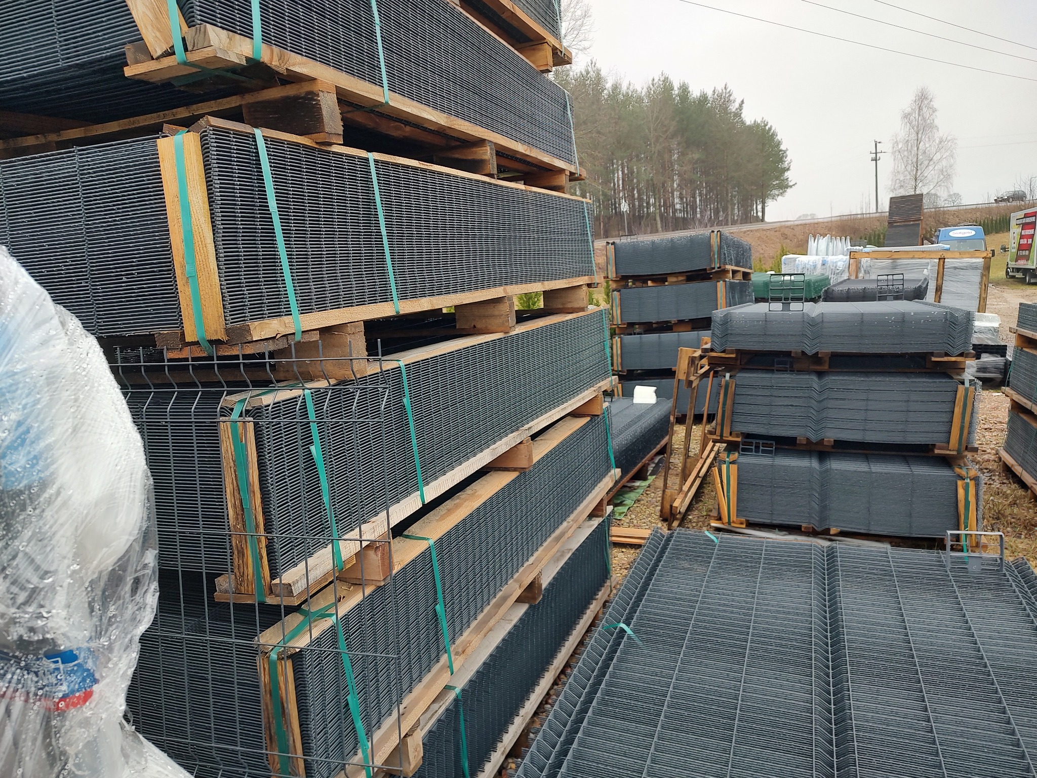 Stosy paneli ogrodzeniowych zgrzewanych, zabezpieczonych taśmami na drewnianych paletach, składowane na zewnątrz w pochmurny dzień.