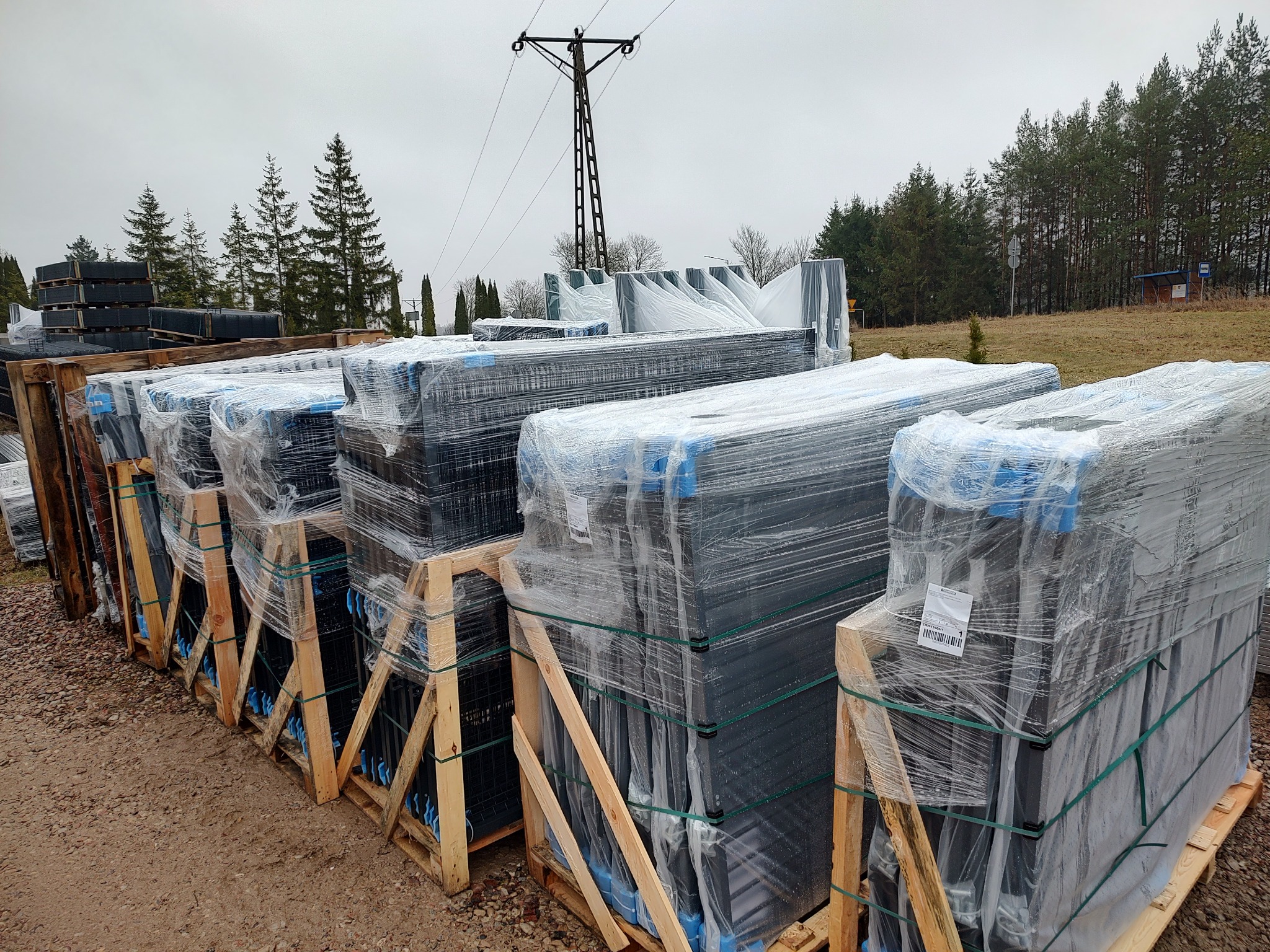 Palety z panelami ogrodzeniowymi, zabezpieczone folią i taśmami, składowane na zewnątrz, widok na słup energetyczny i drzewa w tle.