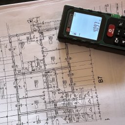 PMKB - Plan architektoniczny domu z naniesionymi wymiarami, obok leży dalmierz laserowy wskazujący 4.097 metra.