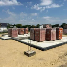 Fundament domu jednorodzinnego we Wrocławiu z przygotowanymi paletami cegieł ceramicznych Termoton, zabezpieczonymi folią, na tle błękitnego nieba z chmurami.