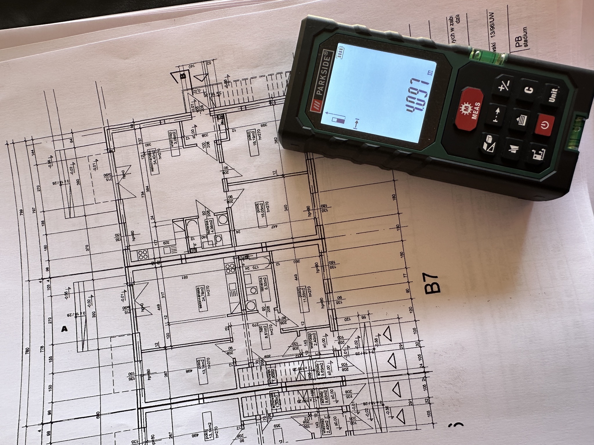 Plan architektoniczny domu z naniesionymi wymiarami, obok leży dalmierz laserowy wskazujący 4.097 metra.