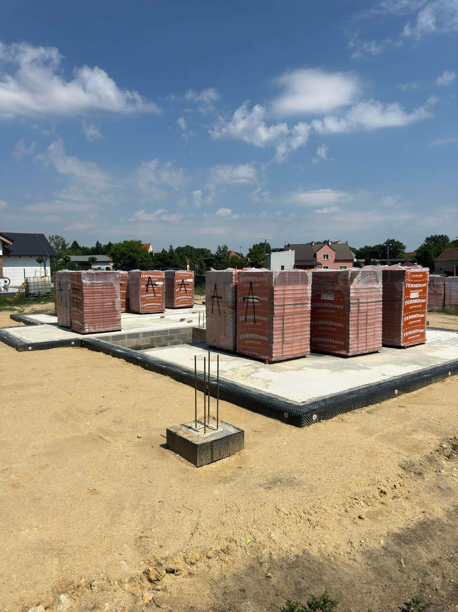 Fundament domu jednorodzinnego we Wrocławiu z przygotowanymi paletami cegieł ceramicznych Termoton, zabezpieczonymi folią, na tle błękitnego nieba z chmurami.