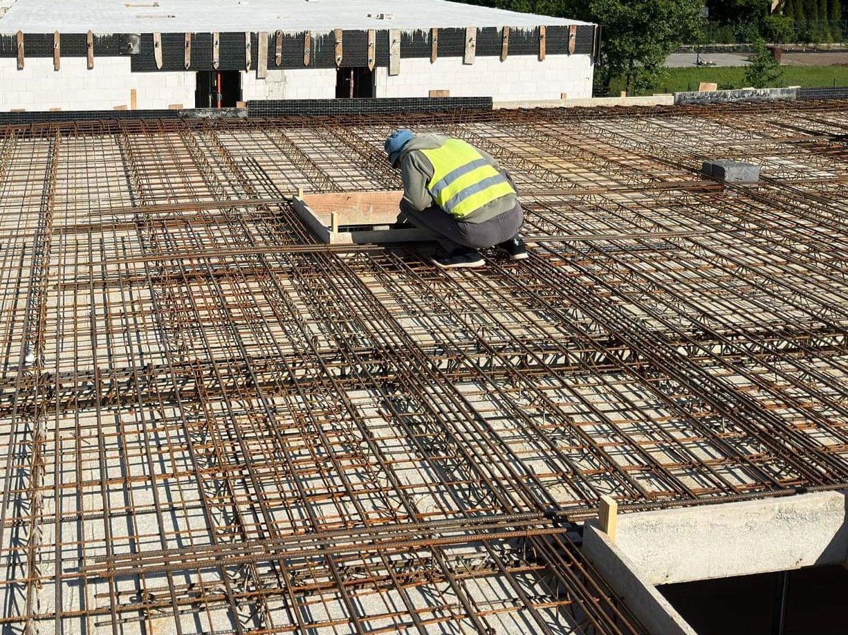 Pracownik w kamizelce odblaskowej i kasku, w kucki, układa zbrojenie na stropie budynku w trakcie budowy, widoczne siatki z prętów stalowych i elementy szalunku.