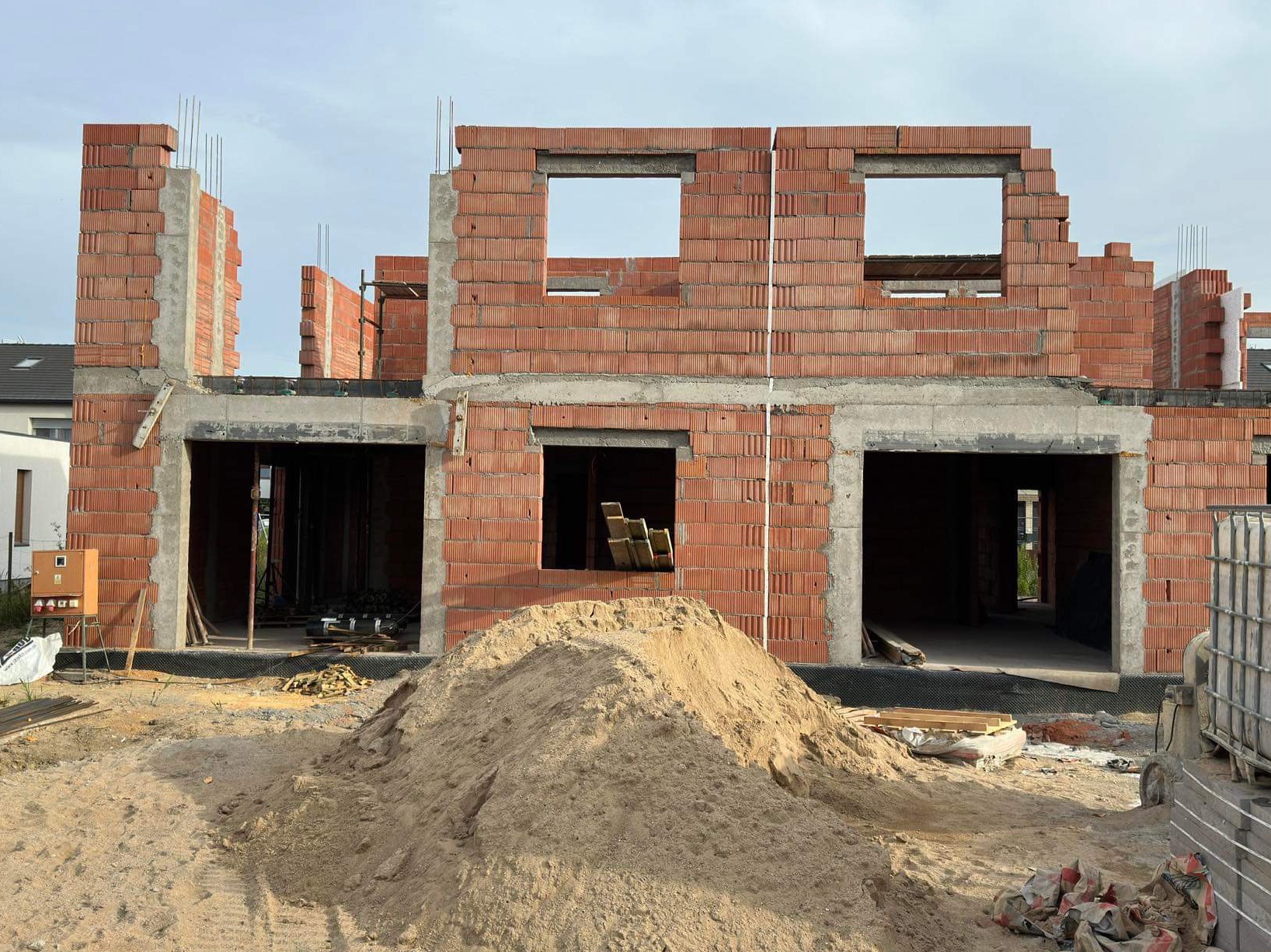 Surowy stan dwukondygnacyjnego domu z czerwonej cegły z widocznymi otworami okiennymi i drzwiowymi, na pierwszym planie hałda piasku, widoczne zbrojenia i elementy konstrukcyjne.
