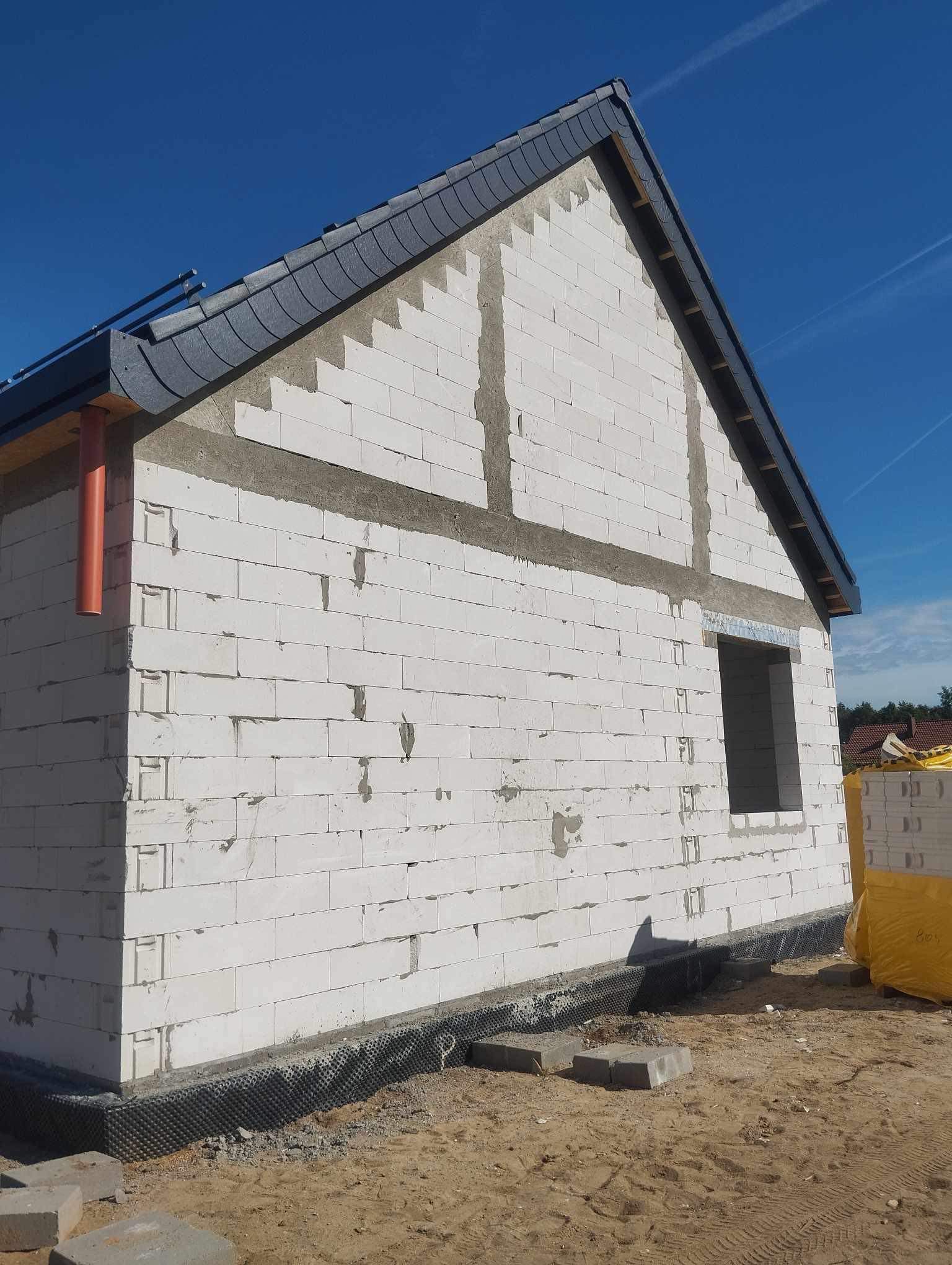 Surowy stan domu jednorodzinnego z białych bloczków, widoczne zbrojenia i okno bez ramy, czarne dachówki na skośnym dachu, teren wokół budynku piaszczysty.