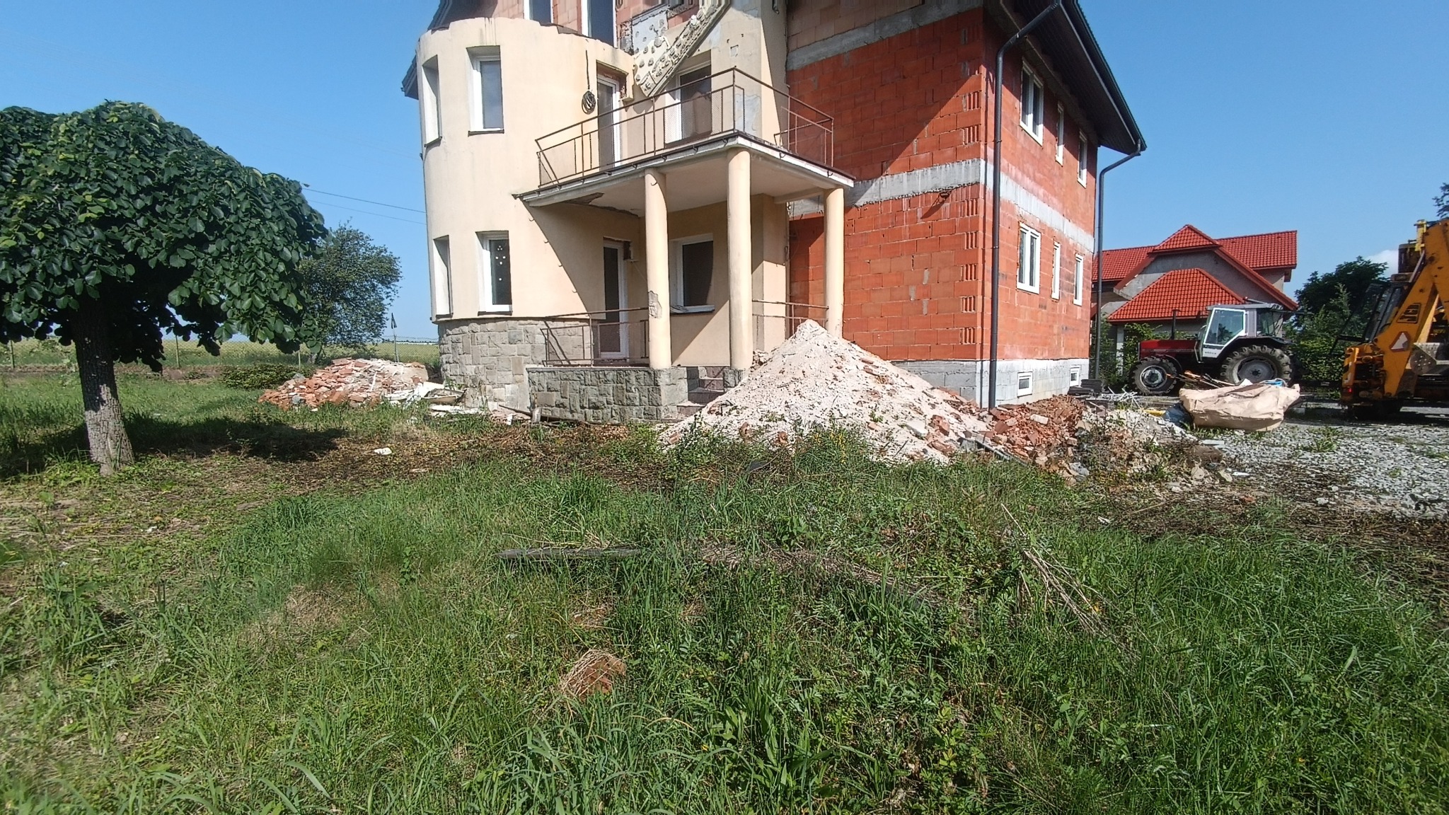 Budynek w trakcie rozbiórki lub przebudowy, widoczne cegły, gruz i maszyny budowlane na zewnątrz, dom z niezakończoną elewacją.