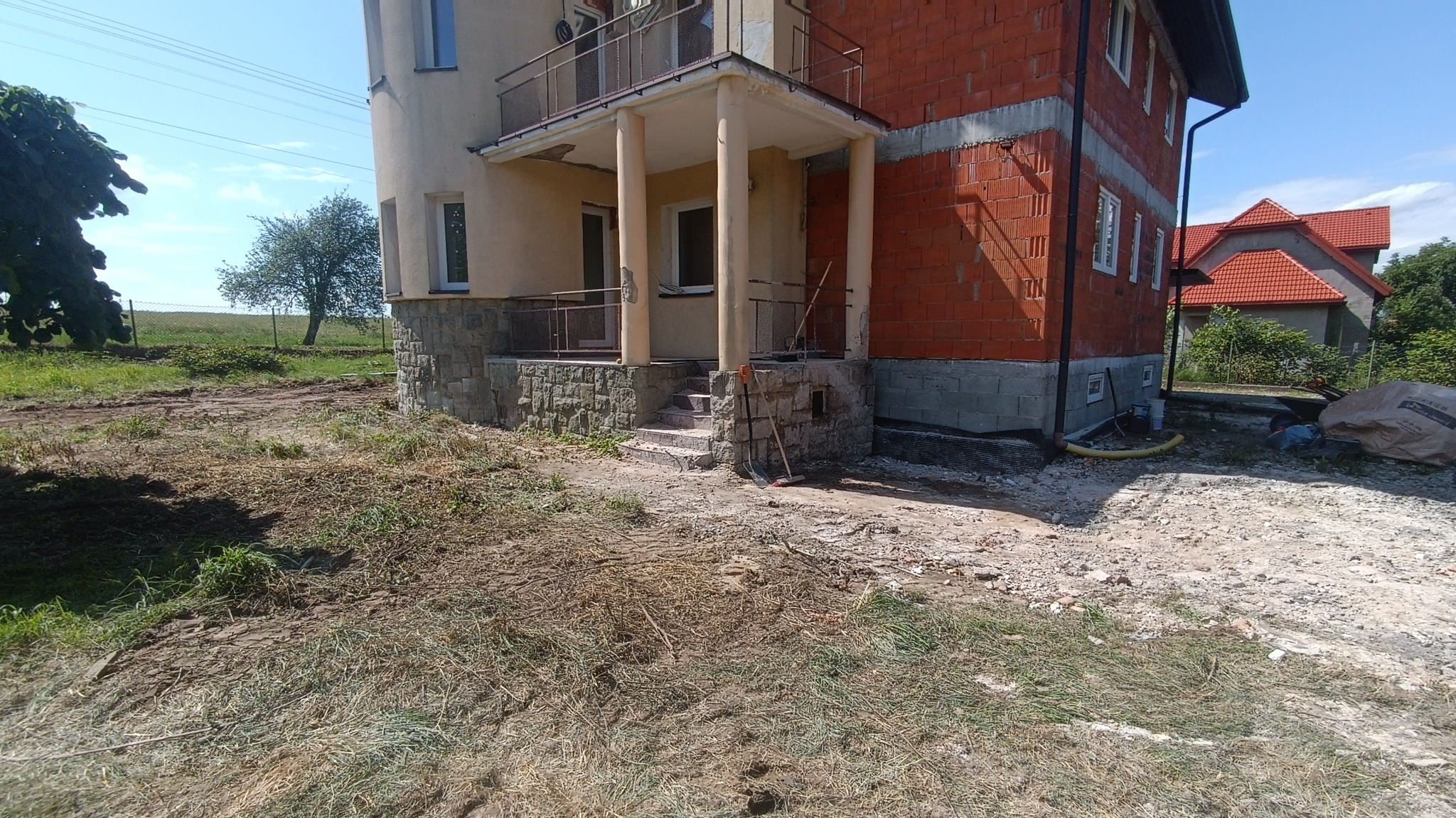Teren wokół domu w trakcie budowy, widoczne świeżo przekopane podłoże, fragmenty trawy, budynek z czerwonej cegły i fragment domu z balkonem, narzędzia oparta o schody.