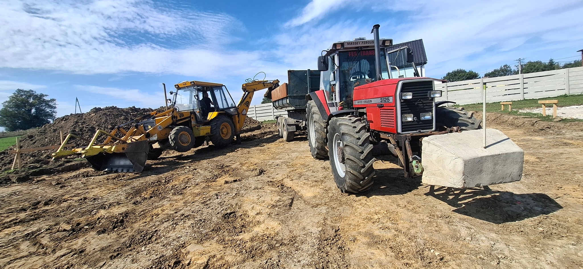 Żółta koparko-ładowarka i czerwony traktor Massey Ferguson na placu budowy z ziemią i betonowym obciążnikiem, w tle błękitne niebo z chmurami.
