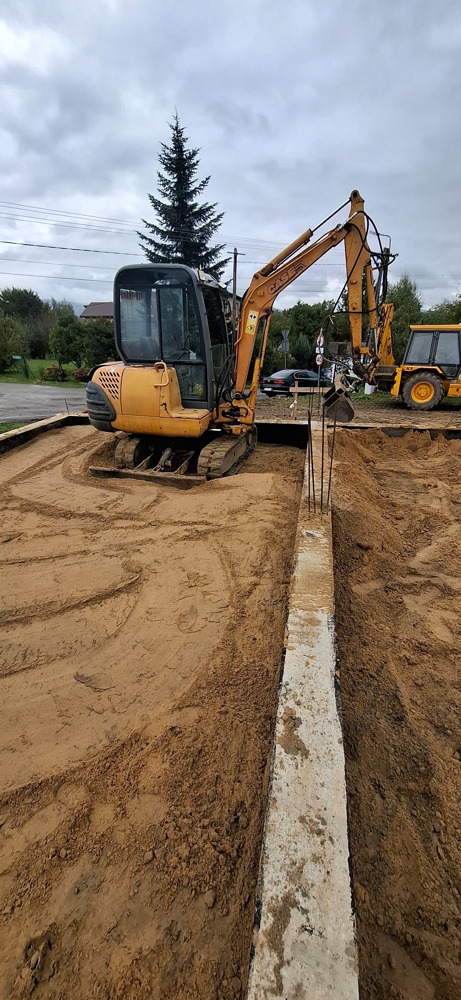 Żółta koparka CASE na placu budowy, obok wykopany rów z betonową ścianą. W tle drzewa i pochmurne niebo. Widoczne ślady opon na piasku.