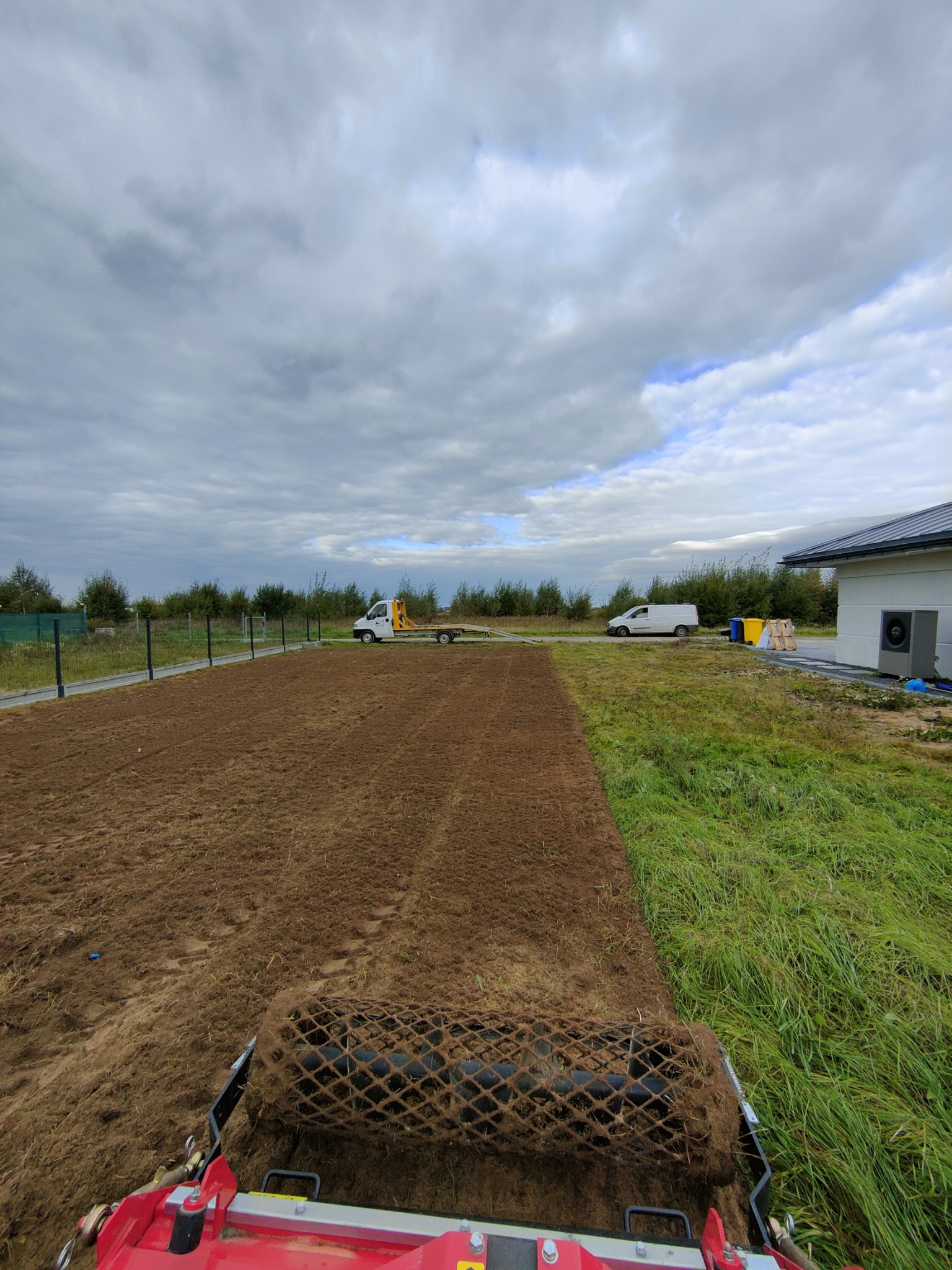 Niwelacja terenu pod trawnik za pomocą wału kolczastego, widok z perspektywy operatora, w tle ogrodzenie, dom i zaparkowane samochody dostawcze