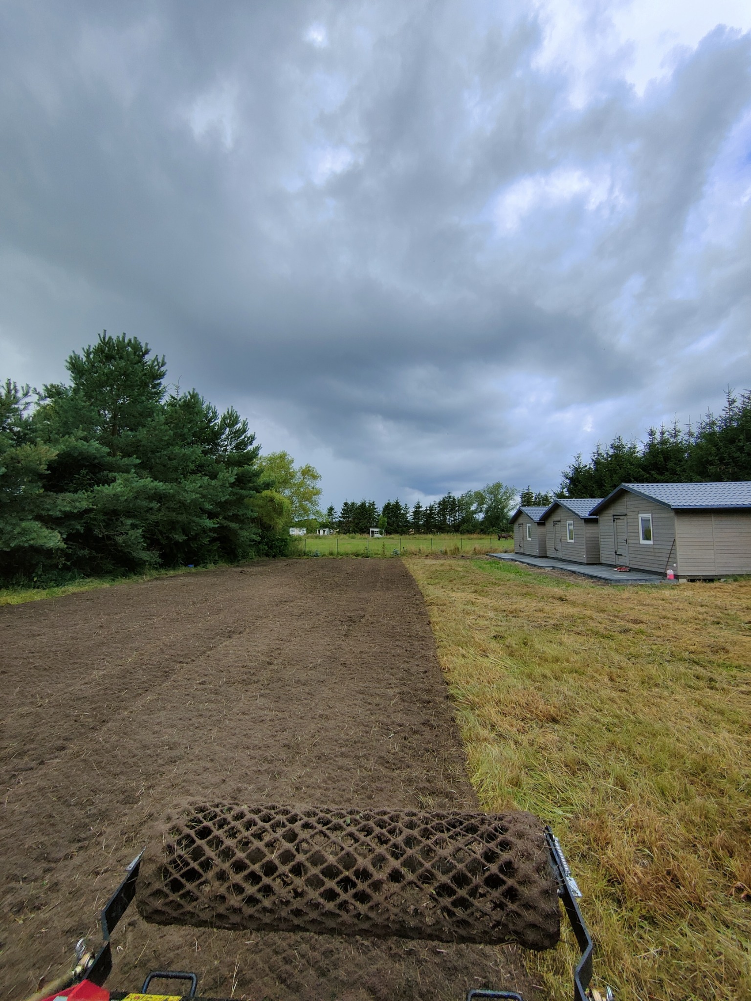 Ciężki walec ugniata świeżo zaorane pole pod zasiew trawy, w tle widoczne budynki gospodarcze i pochmurne niebo.